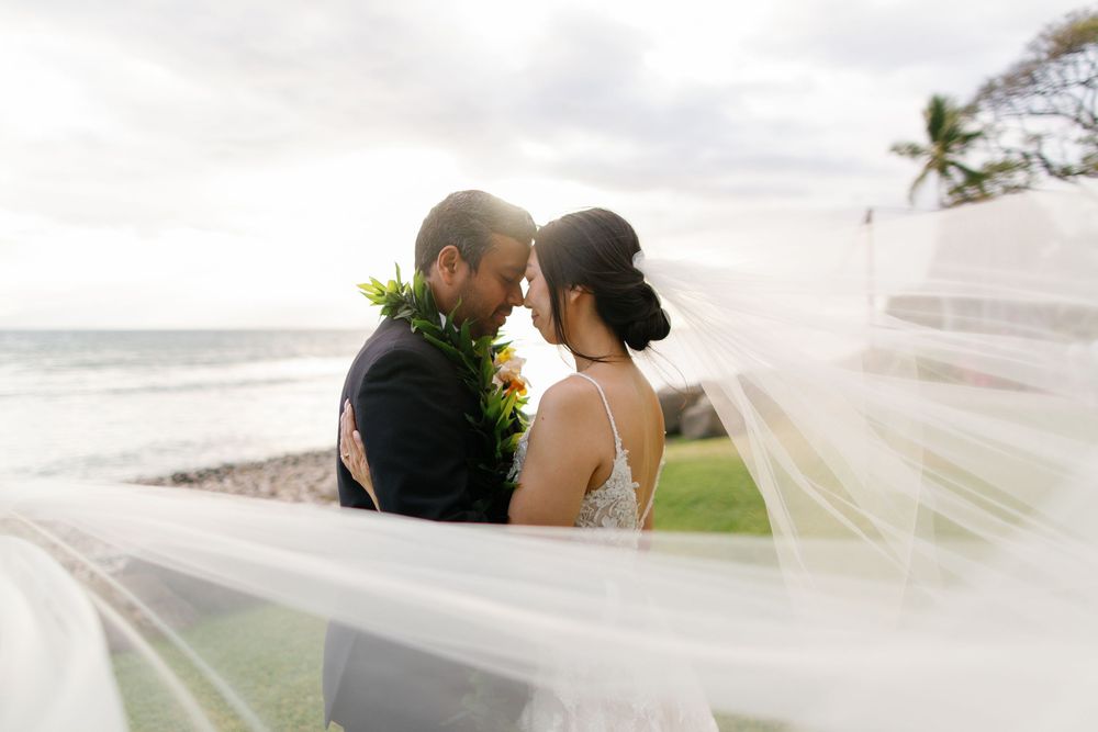 Bride and groom share an intimate moment on a Hawaiian beach, with a flowing veil billowing in the ocean breeze.