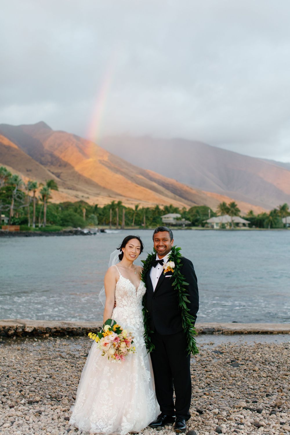 Bride and groom pose by ocean with rainbow and mountains in Hawaii wedding portrait, Olowalu Plantation House wedding.