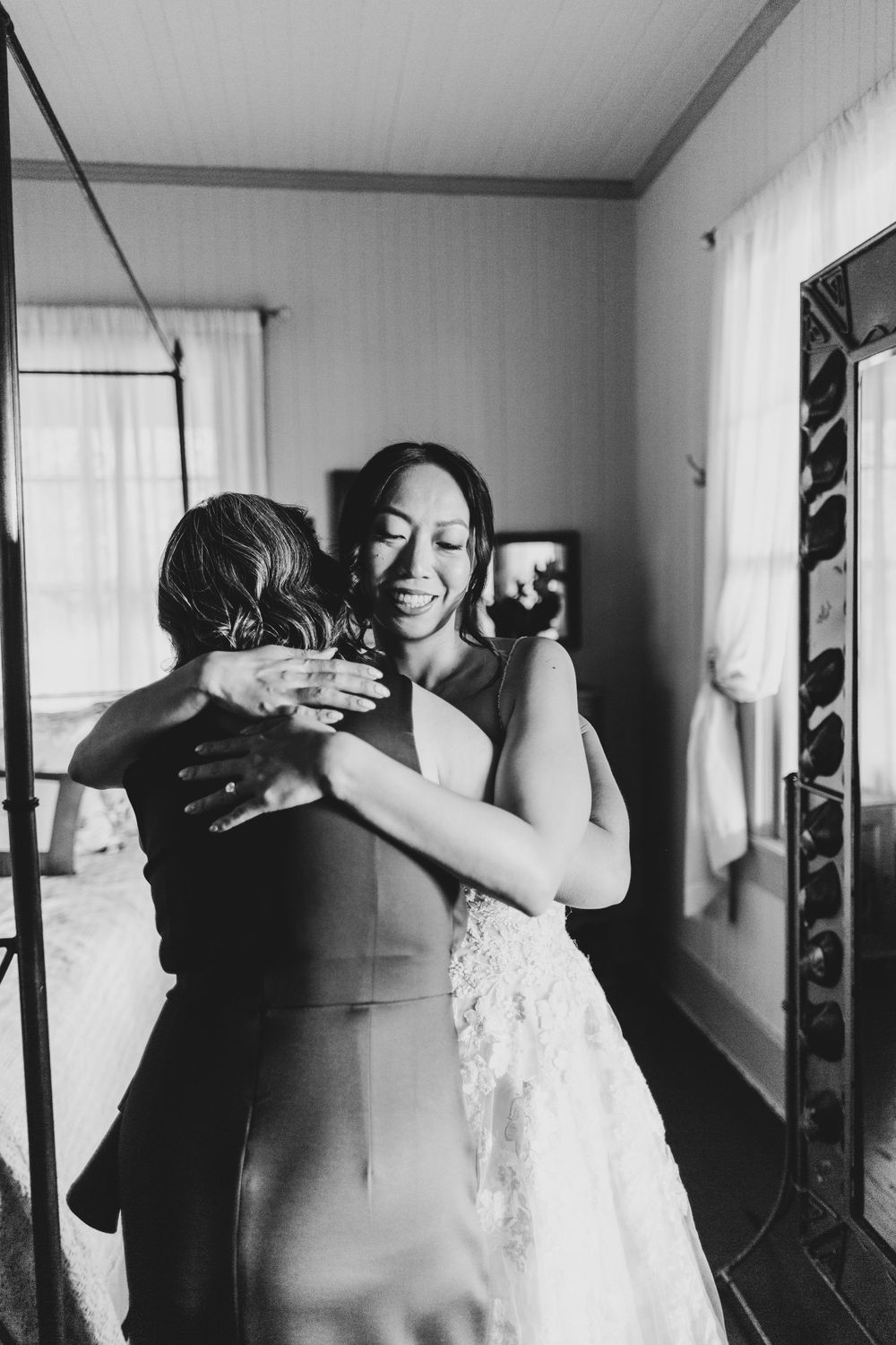 Black and white photo of a bride in a lace gown sharing an emotional hug with a guest during wedding preparations.