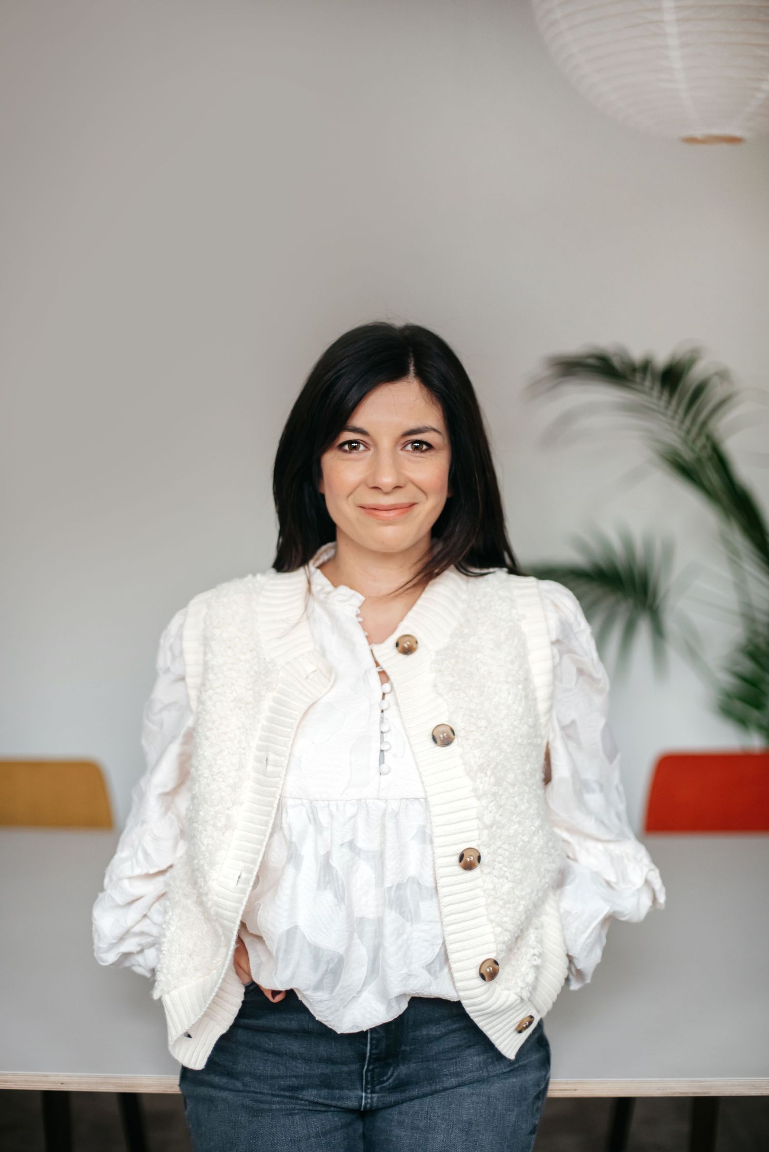 A person wearing a white button-up cardigan and white blouse stands casually against a light wall with a palm plant in background.