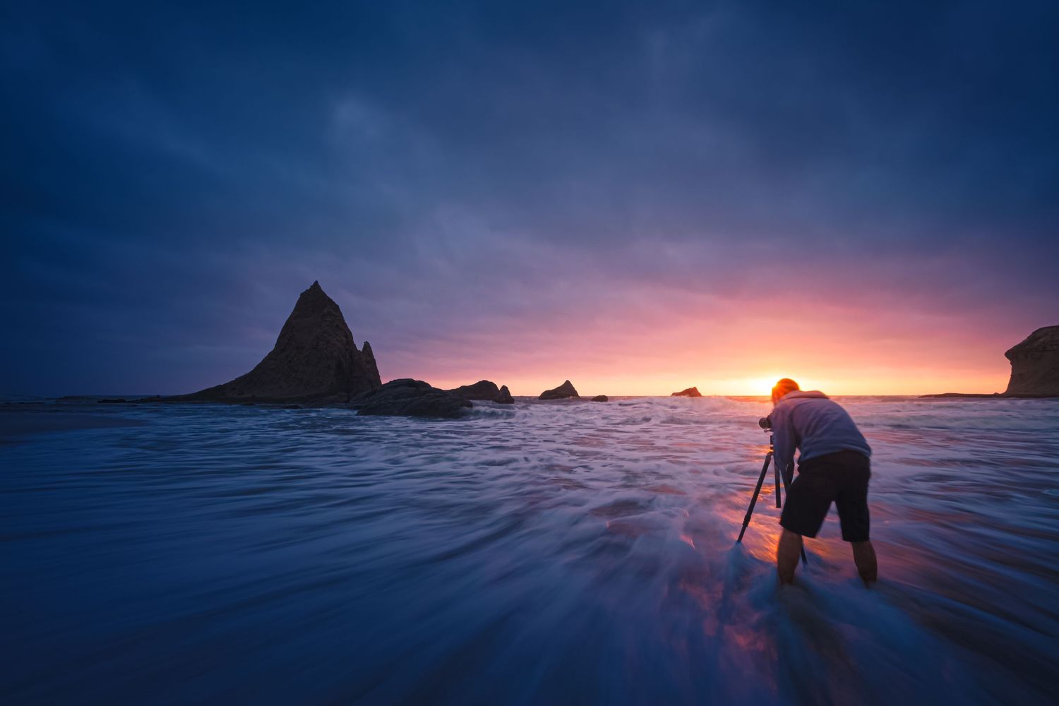 Silhouette of photographer capturing dramatic coastal sunset among rock formations at low tide.