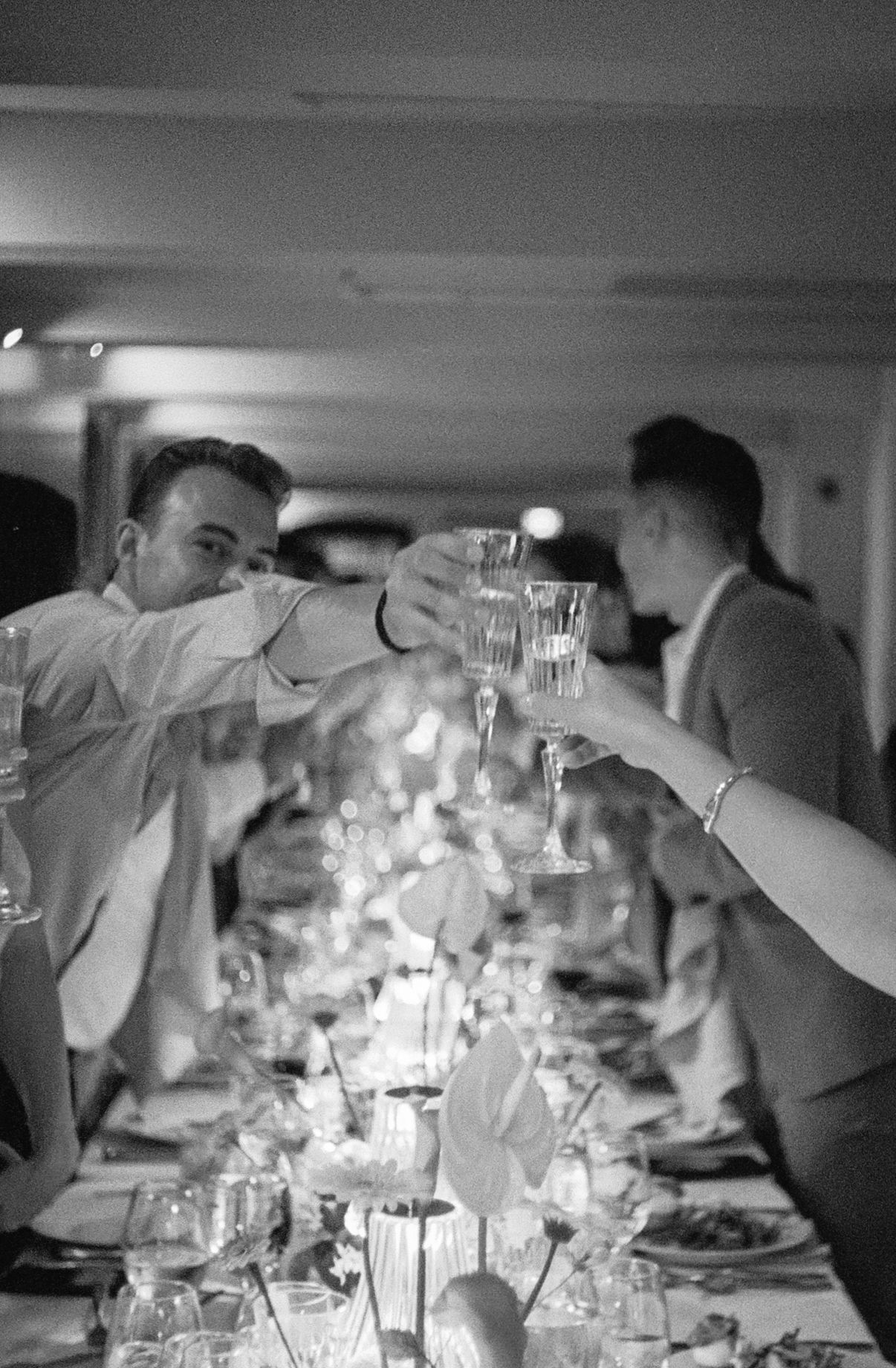 People raise wine glasses in a toast across a candlelit dining table during an elegant black and white dinner party.