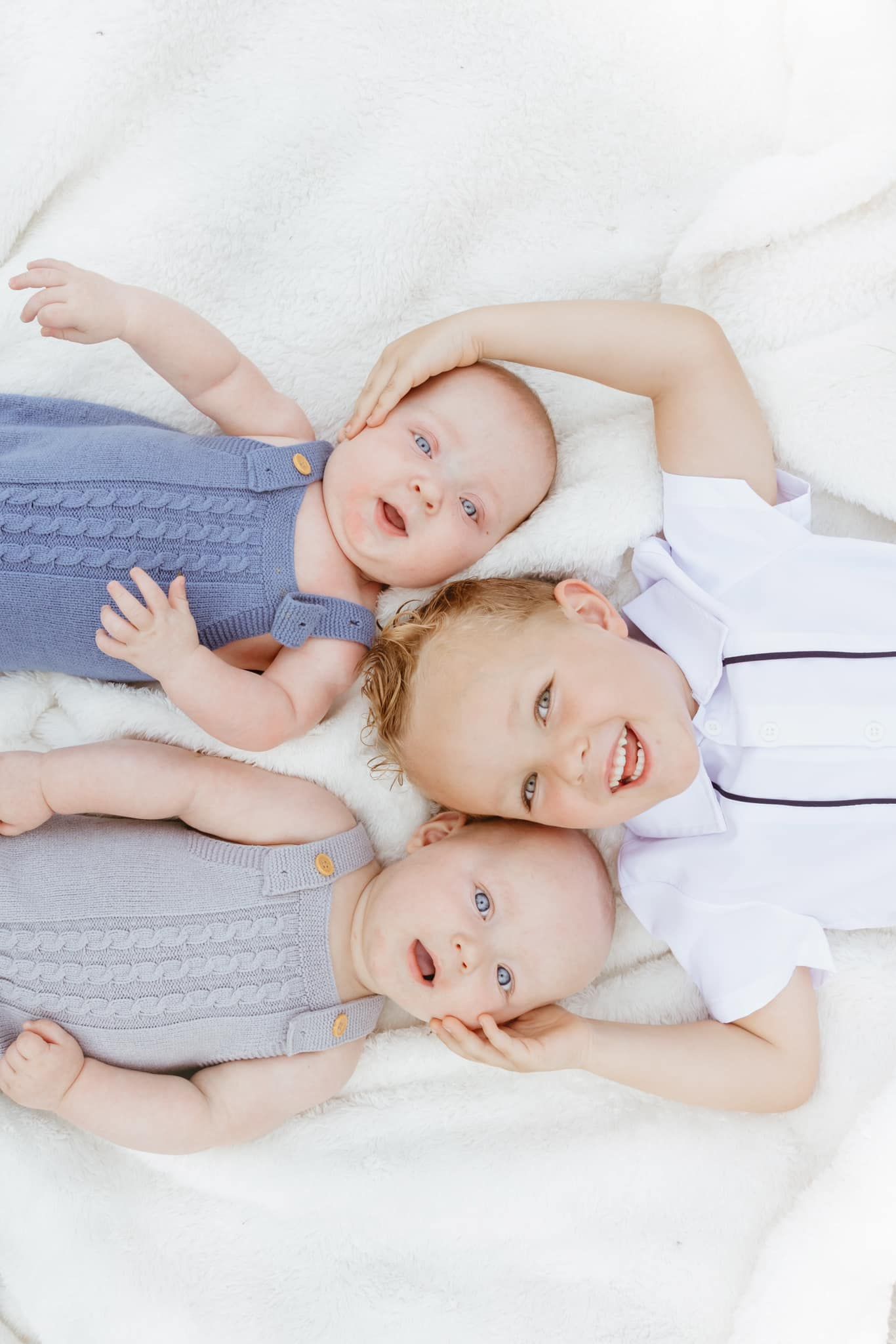 Three young siblings in blue and white outfits lying together on white bedding.
