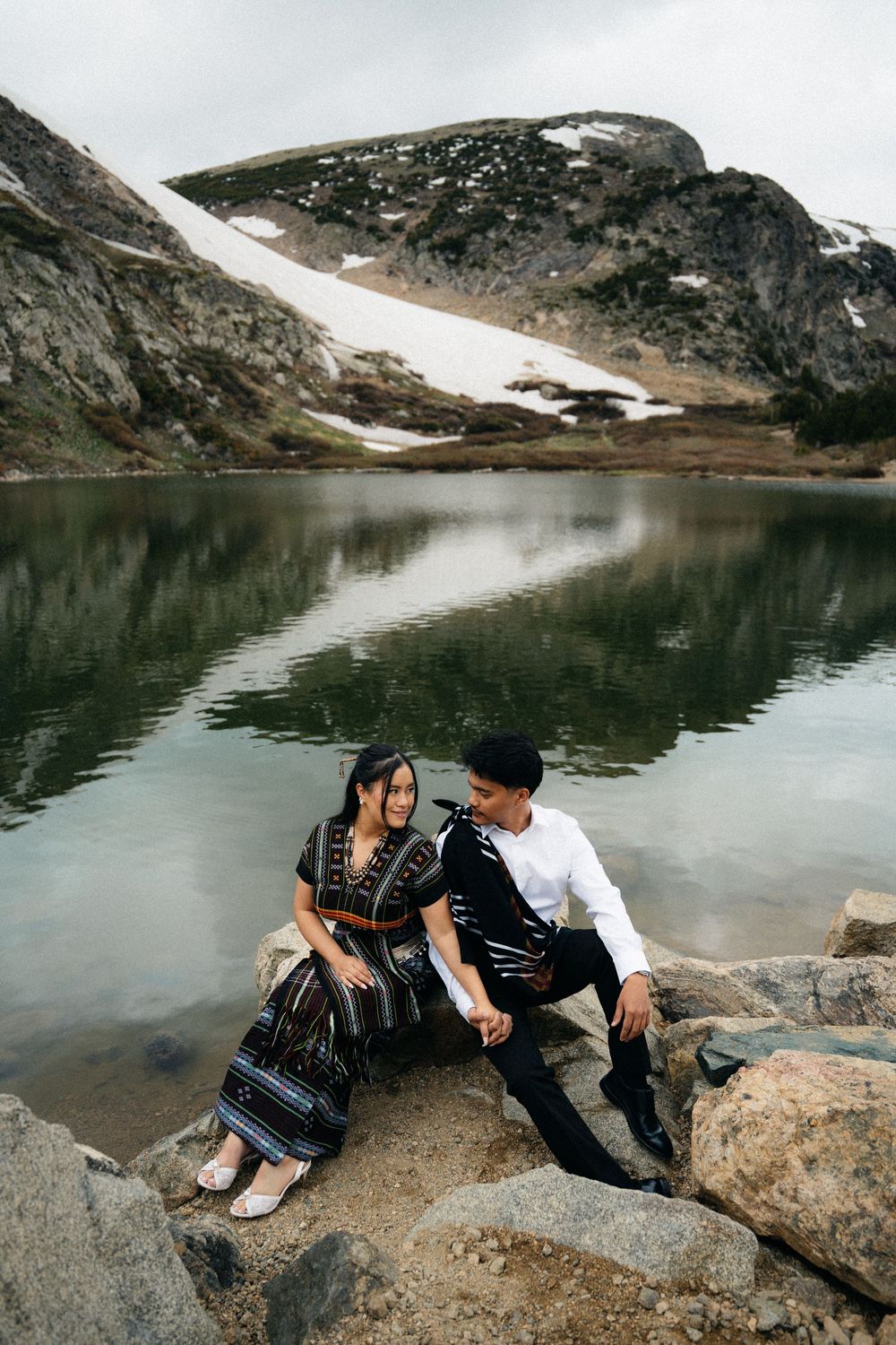 Couple sitting on rocks by a serene mountain lake with snow-capped peaks, wearing elegant traditional attire.