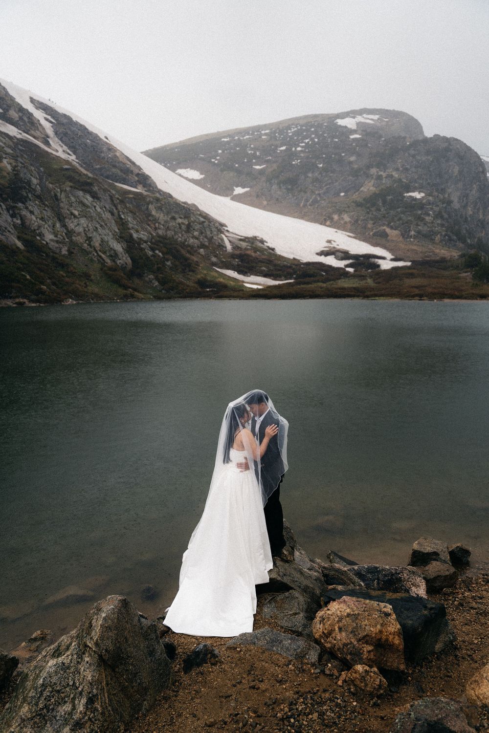 Bride and groom kissing under veil by a mountain lake with snow-capped peaks in the background.