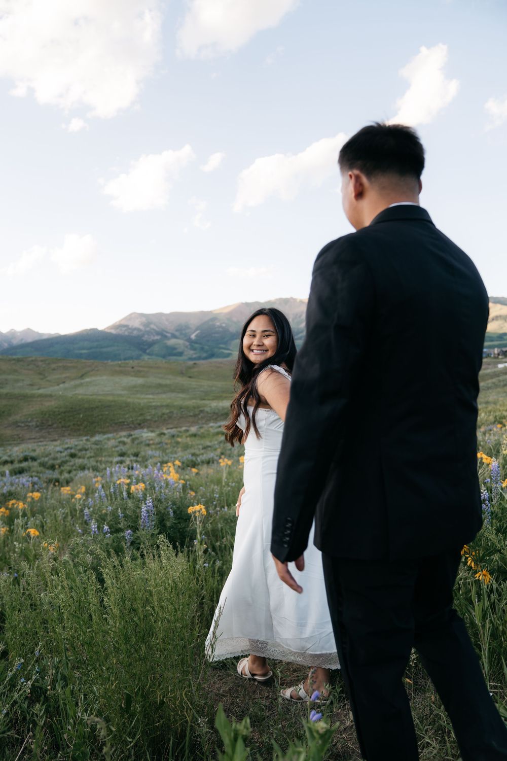 Couple in wedding attire standing in a wildflower meadow with mountain views, bride smiling back at groom.