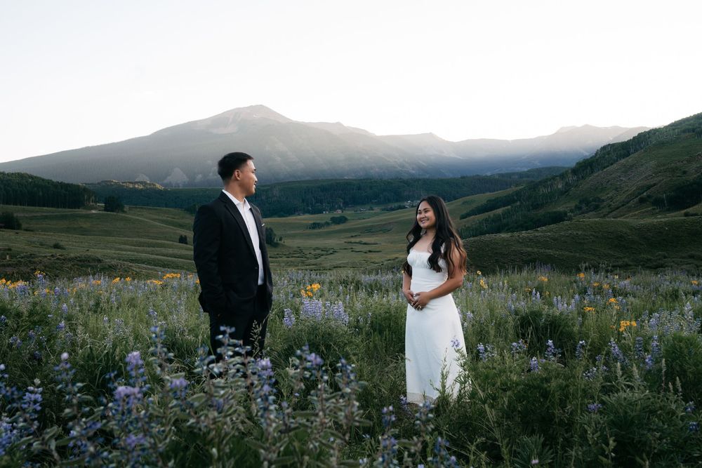 Couple standing in a wildflower meadow with mountain backdrop at sunset, man in black suit, woman in white dress.