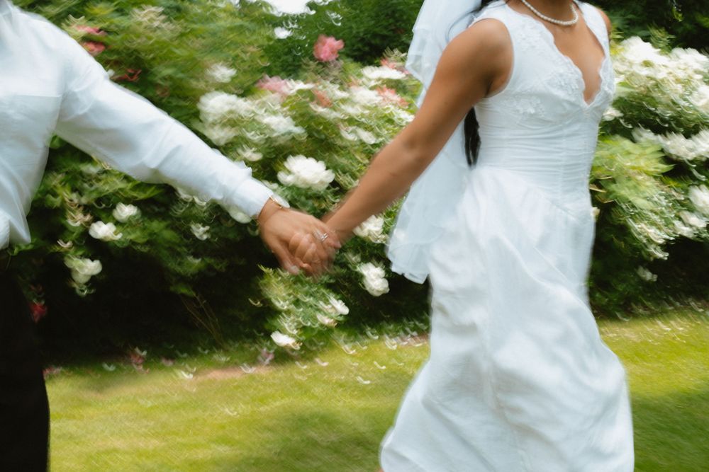 Bride and groom holding hands running through garden with white flowering bushes in background.
