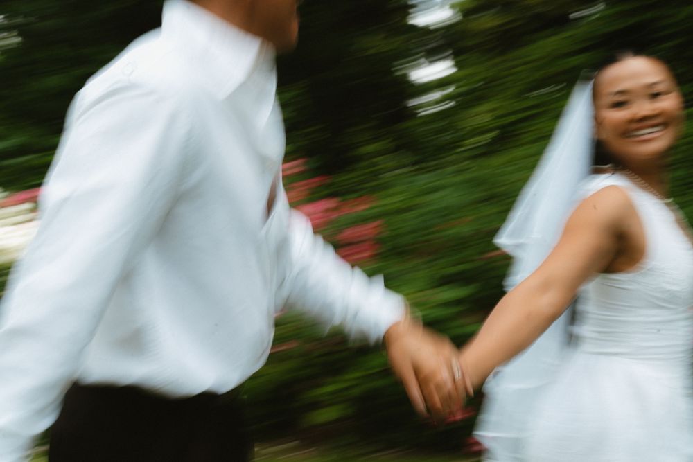 Smiling bride in white veil holding groom's hand while running through lush green garden.