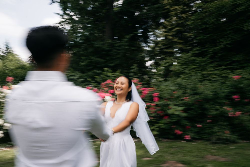 Bride in white dress and veil laughing joyfully outdoors with pink flowers and lush green trees in background.