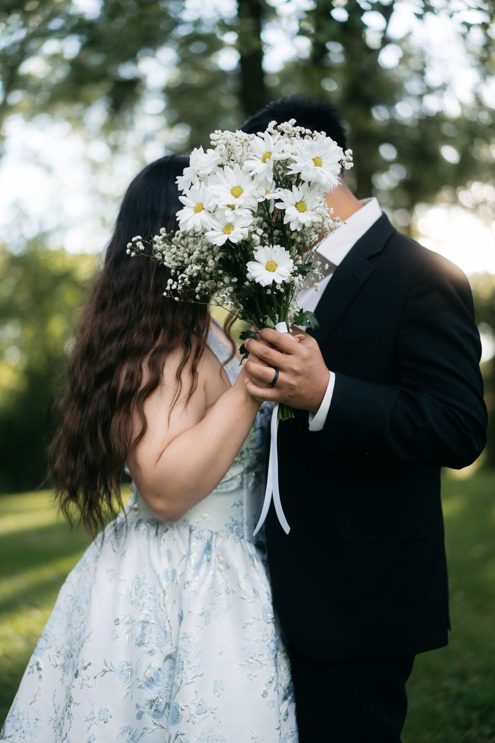 Couple hiding behind white daisy bouquet while embracing outdoors in sunlit green park during engagement shoot.