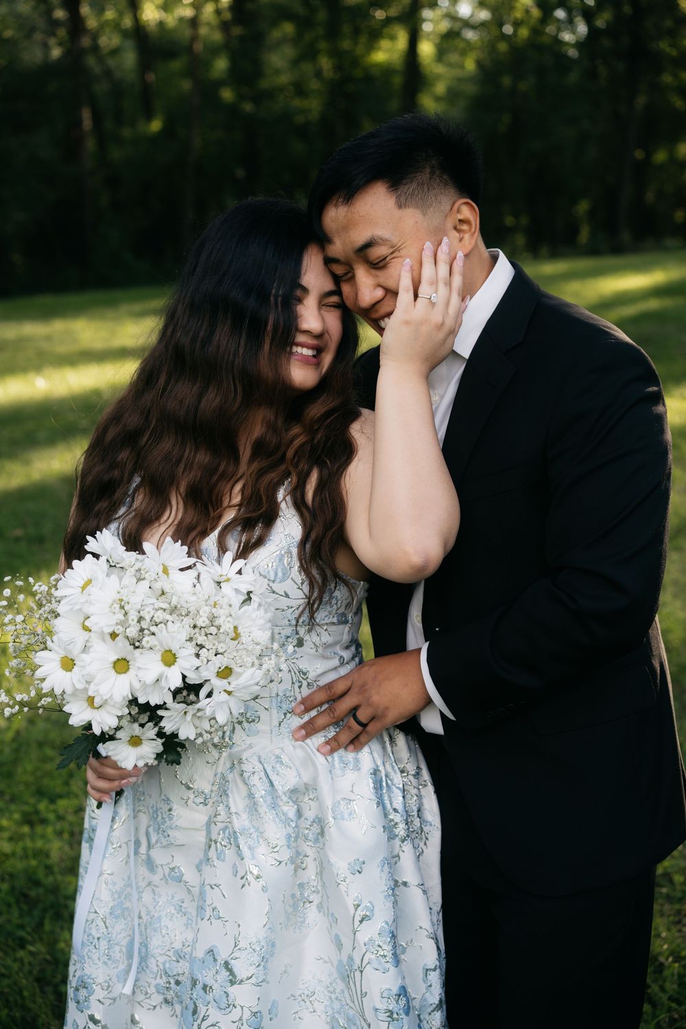 Bride in floral dress tenderly touches grooms face while holding daisy bouquet during golden hour park session.