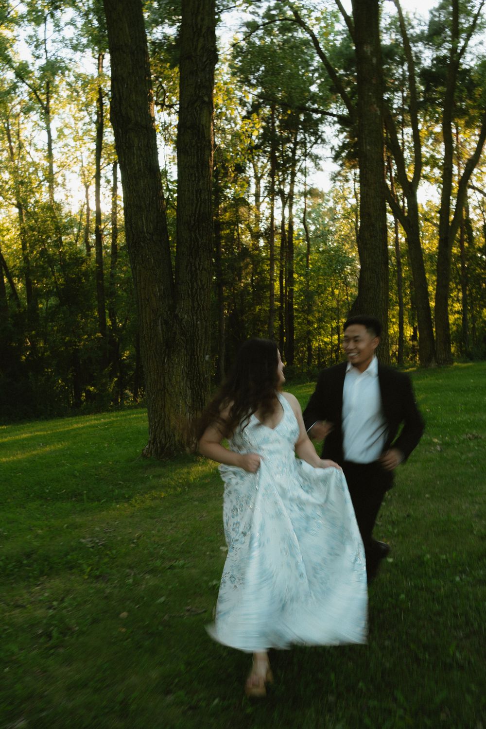 Joyful couple running and laughing together through sunlit forest during outdoor engagement or wedding photo session.