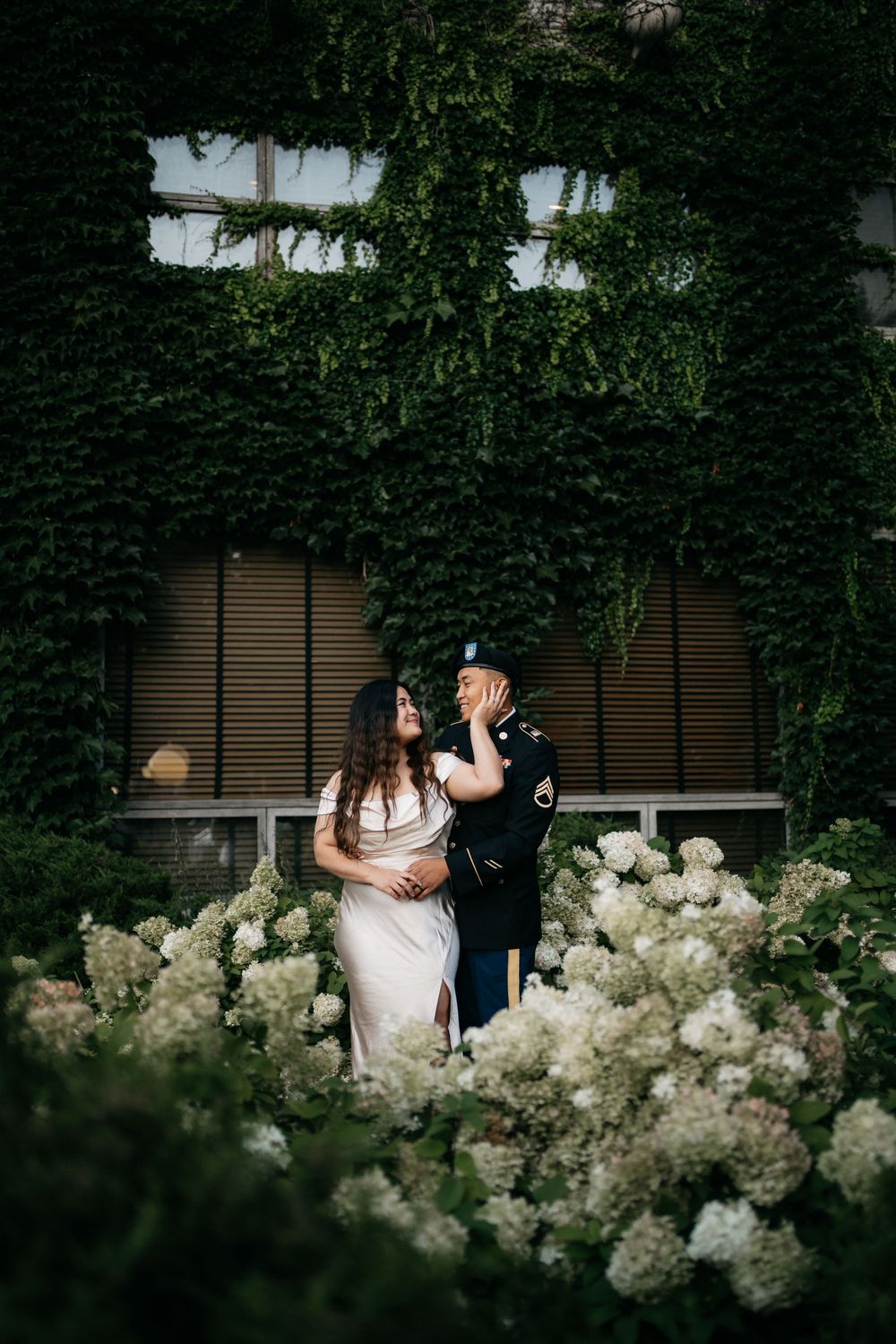 Couple in wedding attire standing romantically among white hydrangeas against ivy-covered wall in urban garden setting.