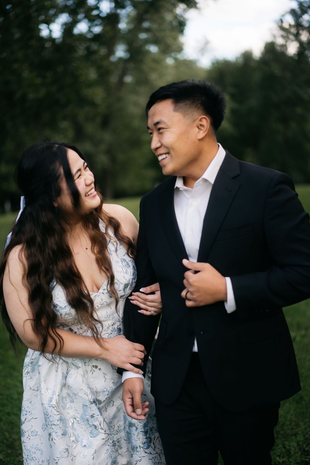 Happy couple in elegant attire laughing together during engagement photo session in a lush green park.