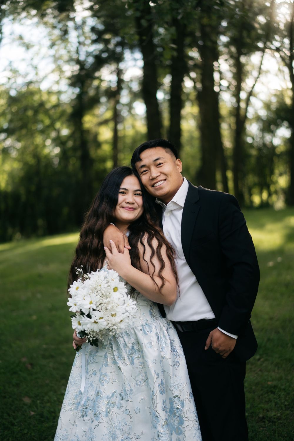 Couple embracing in sunny park, woman in floral dress holding white daisy bouquet, man in black suit smiling.