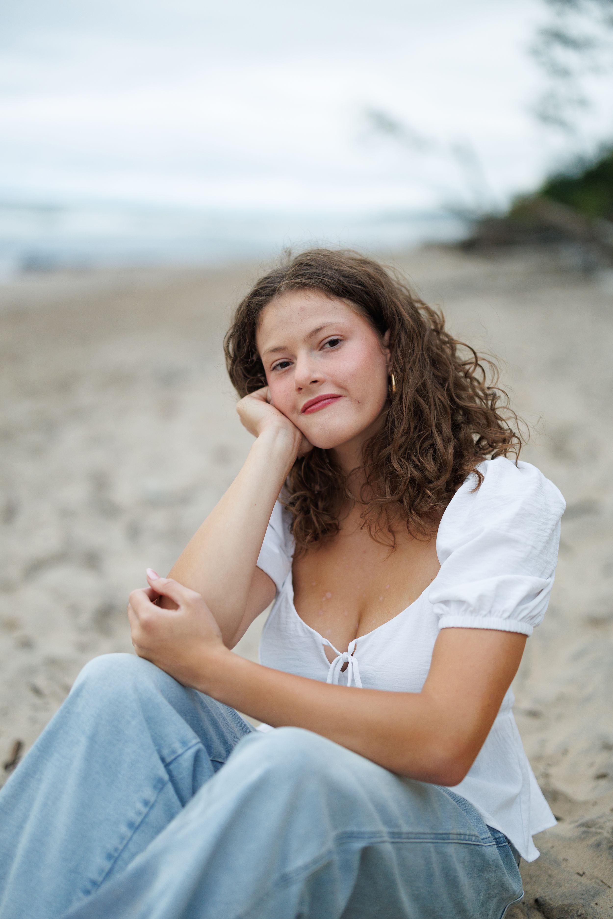 Woman in white top and jeans sits relaxed on a beach with cloudy sky and shoreline in background.