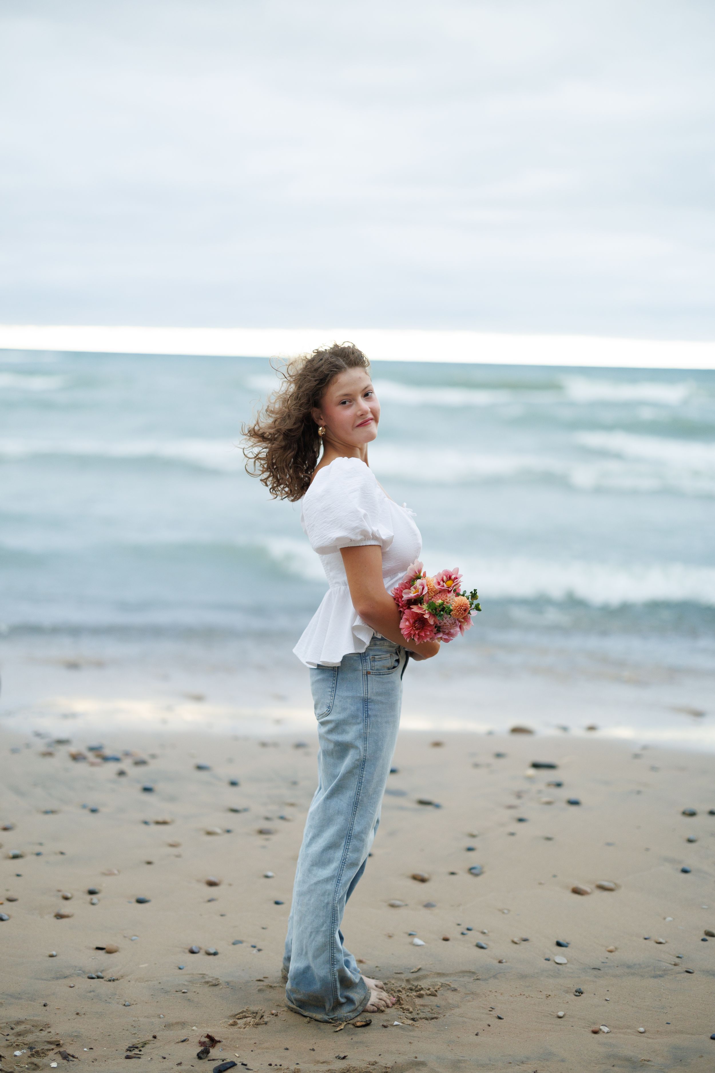 A child in light denim jeans and white shirt stands on beach holding flowers while looking at ocean waves.