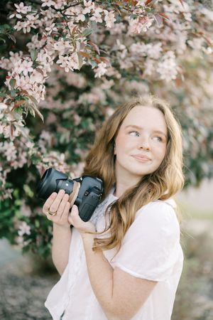 A photographer holds a professional camera under blooming spring cherry blossoms.