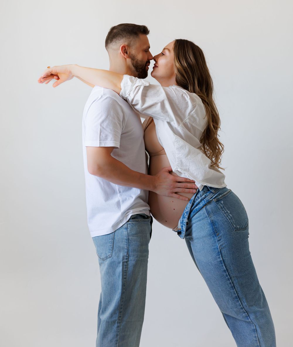 Pregnant couple in white shirts and jeans sharing a tender moment, man holding woman's baby bump against white background.