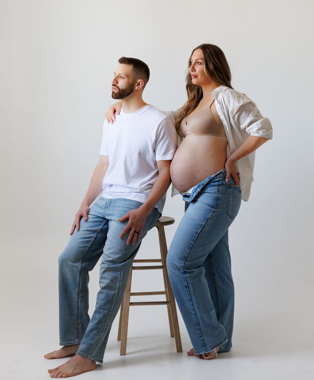 Pregnant couple posing in a studio, man sitting on stool in white tee and jeans, woman standing beside him showing baby bump.