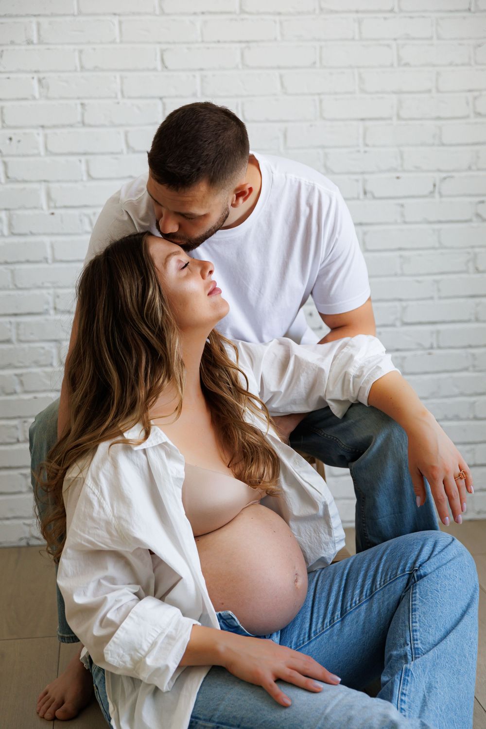 A man tenderly kisses a pregnant woman's forehead as she sits relaxed, both wearing casual white shirts and jeans.