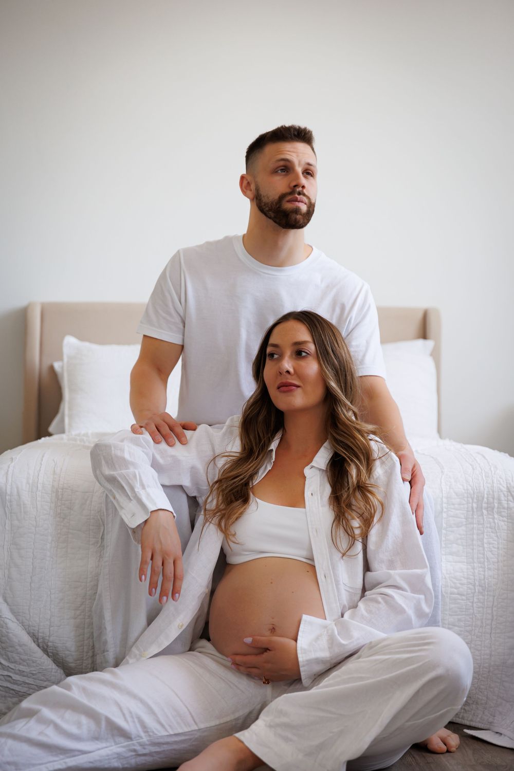 Pregnant woman in white outfit sits on floor with partner behind her, both posing for a maternity photo in a bedroom.