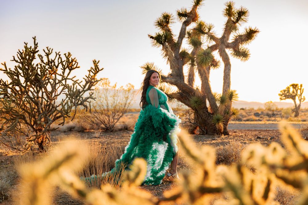 Woman in green gown poses among Joshua trees and cacti in golden desert light.