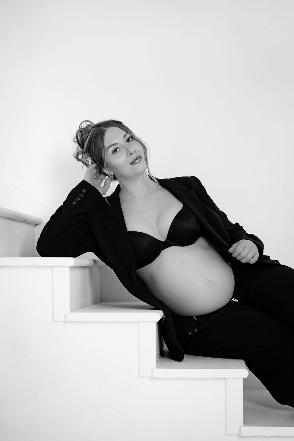 Black and white portrait of a pregnant woman in lingerie and blazer sitting gracefully on white stairs.