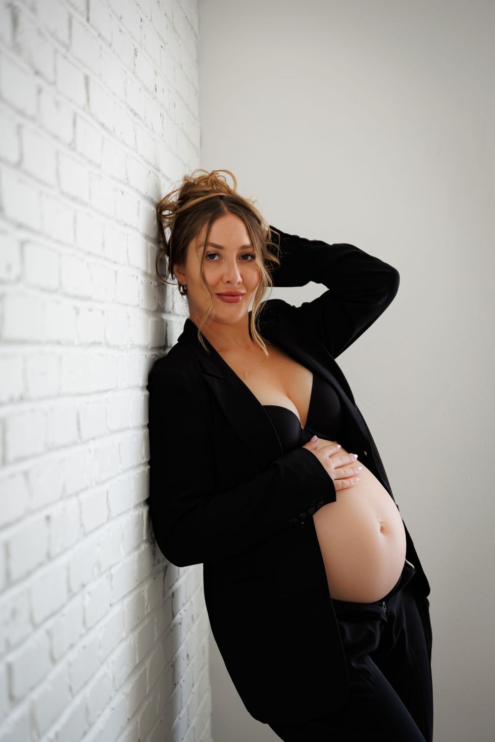 Pregnant woman in black blazer leaning against white brick wall, cradling baby bump in stylish maternity photo shoot.
