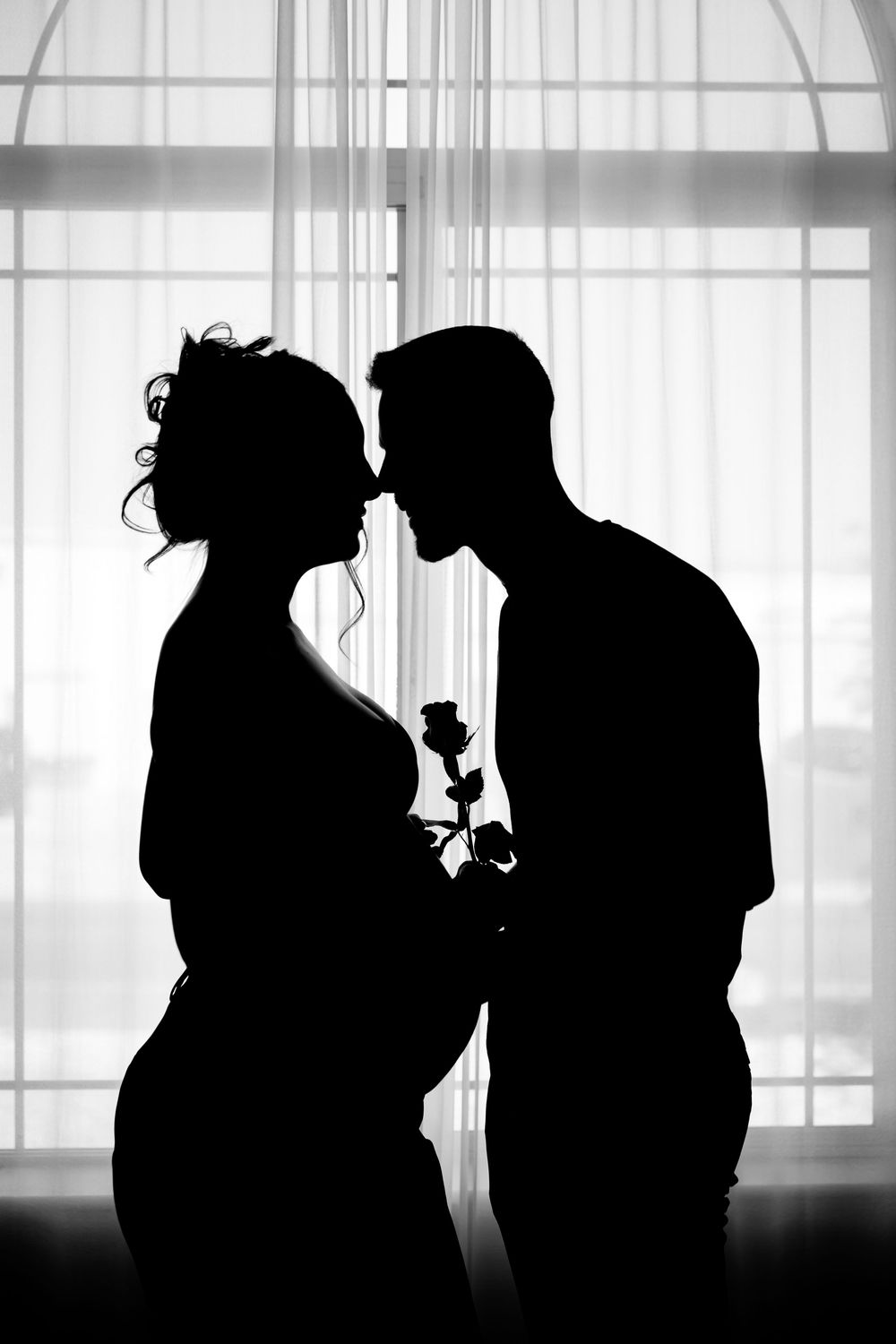 Black and white silhouette of a romantic couple kissing by a window, man holding a rose.