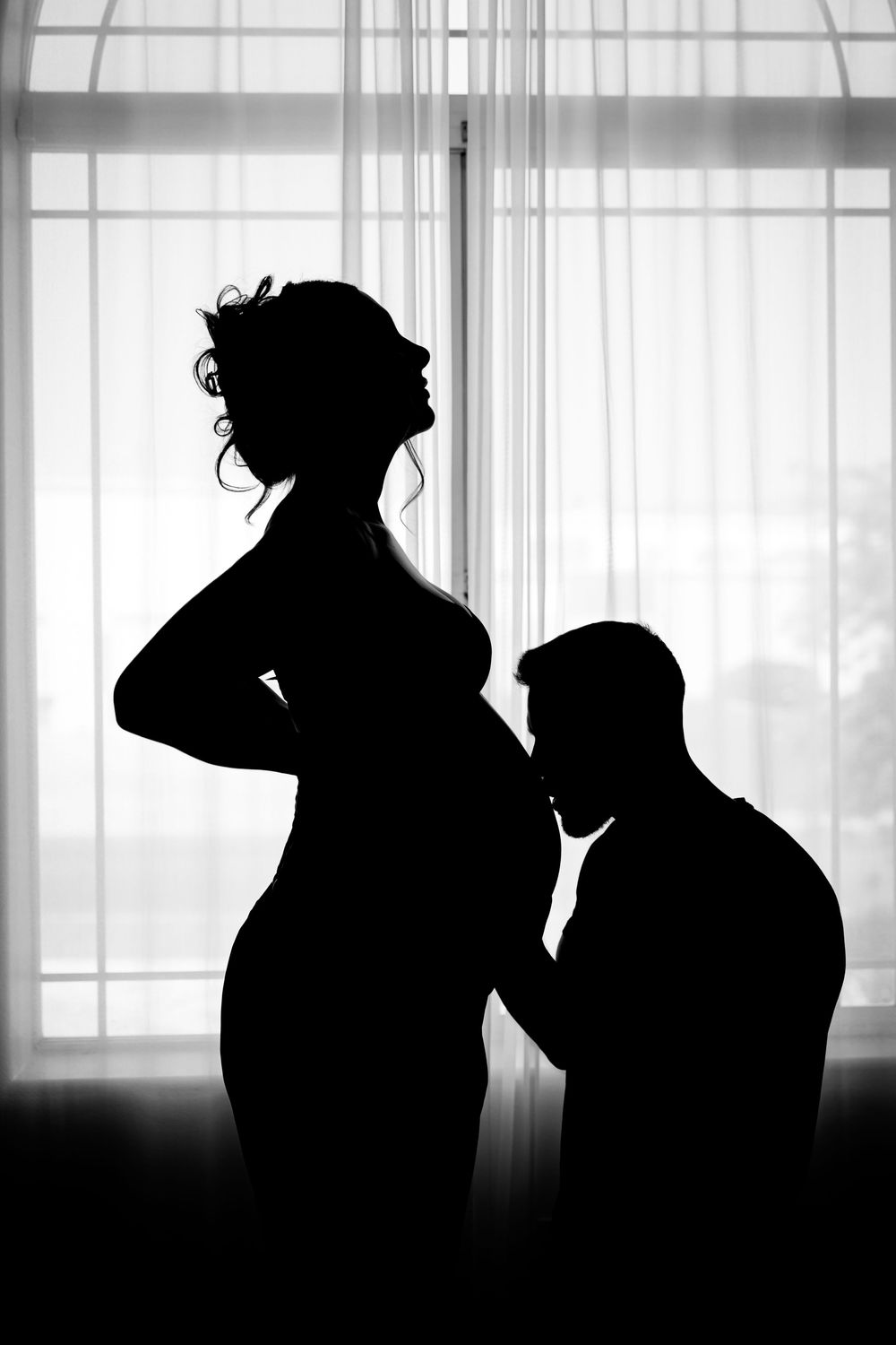 Silhouette of pregnant woman and partner by window in black and white maternity photo.