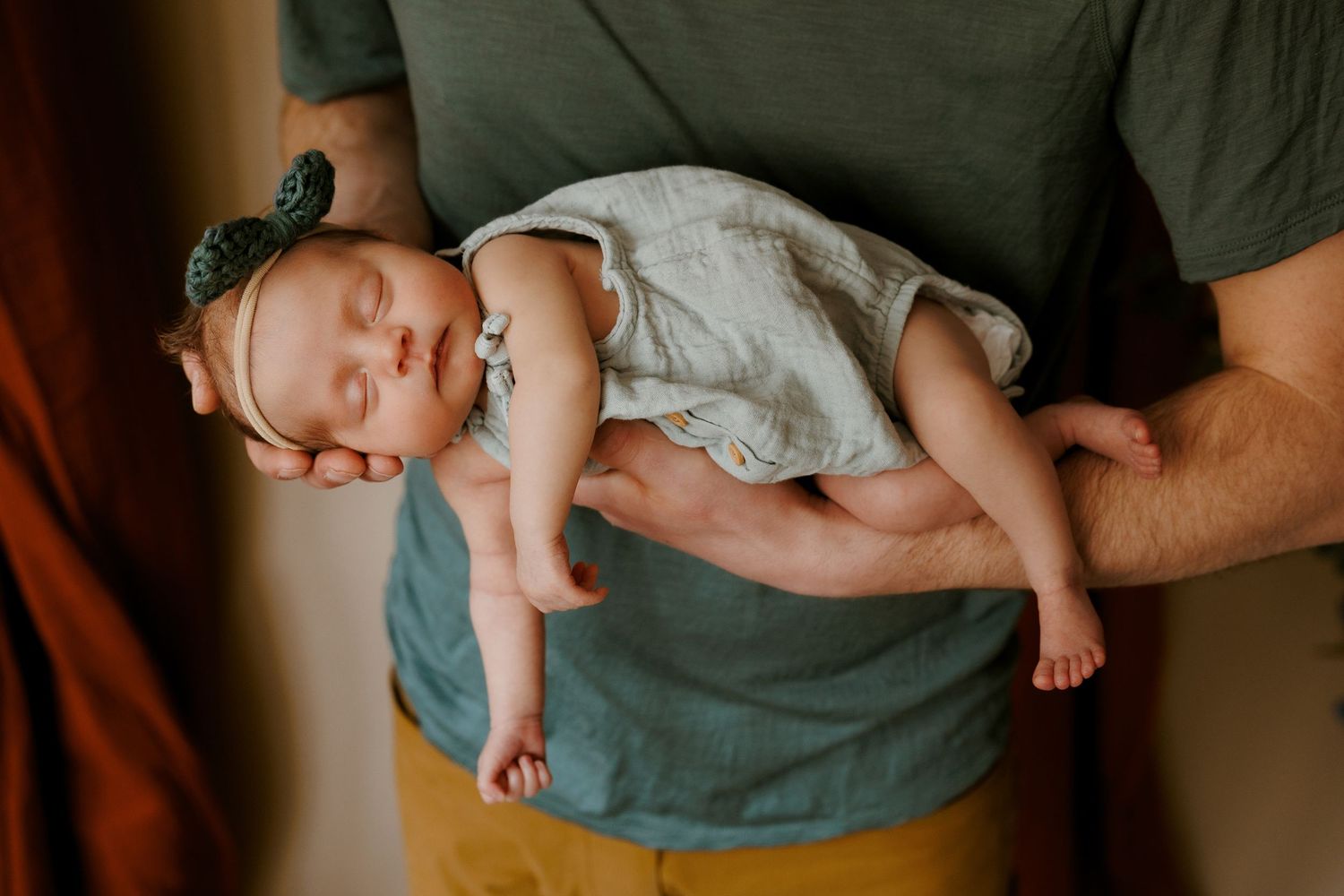 A father gently cradles his sleeping newborn baby dressed in a floral headband.