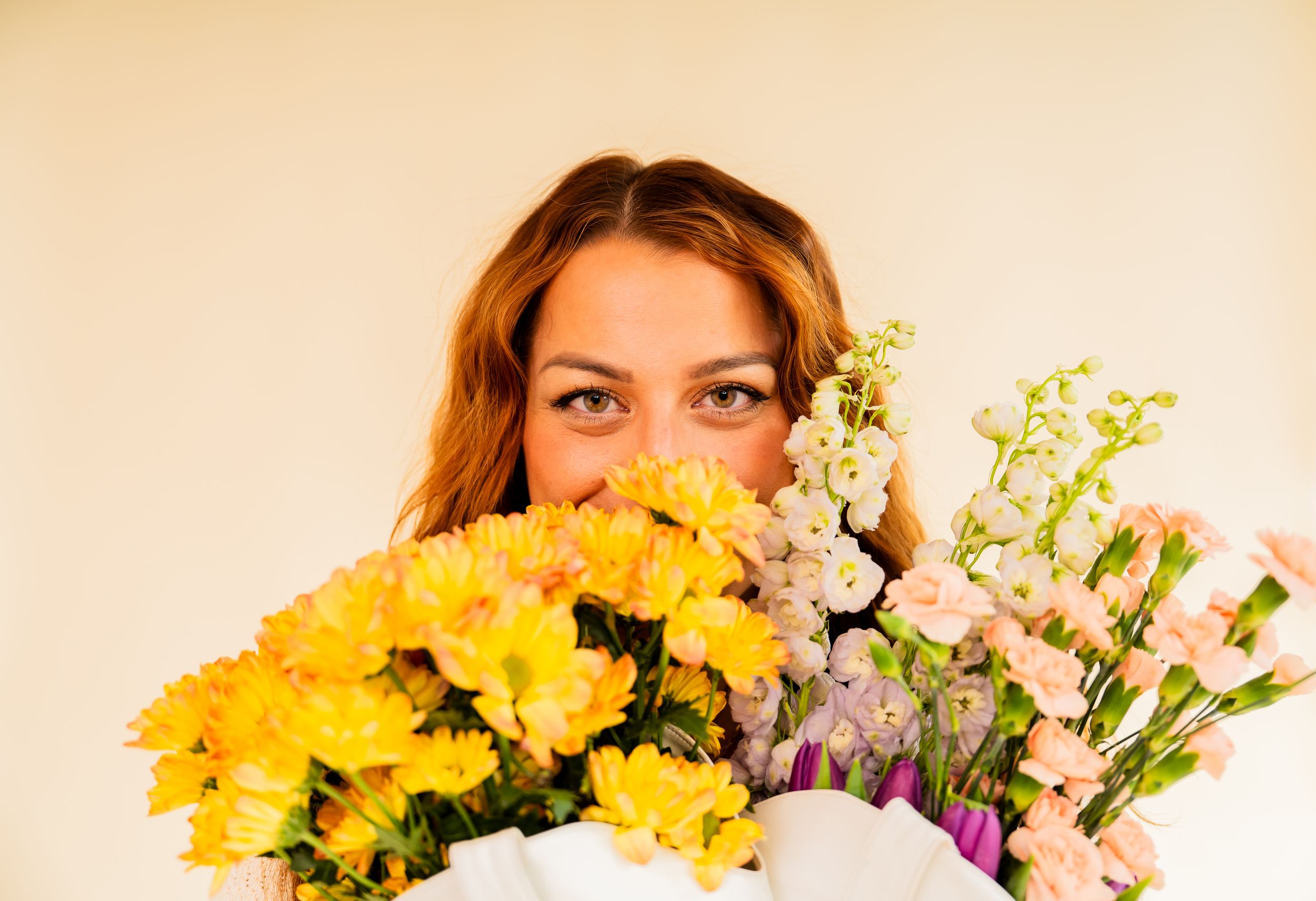 Woman hiding behind colorful bouquet of yellow and pink flowers, with expressive eyes peeking over blooms.
