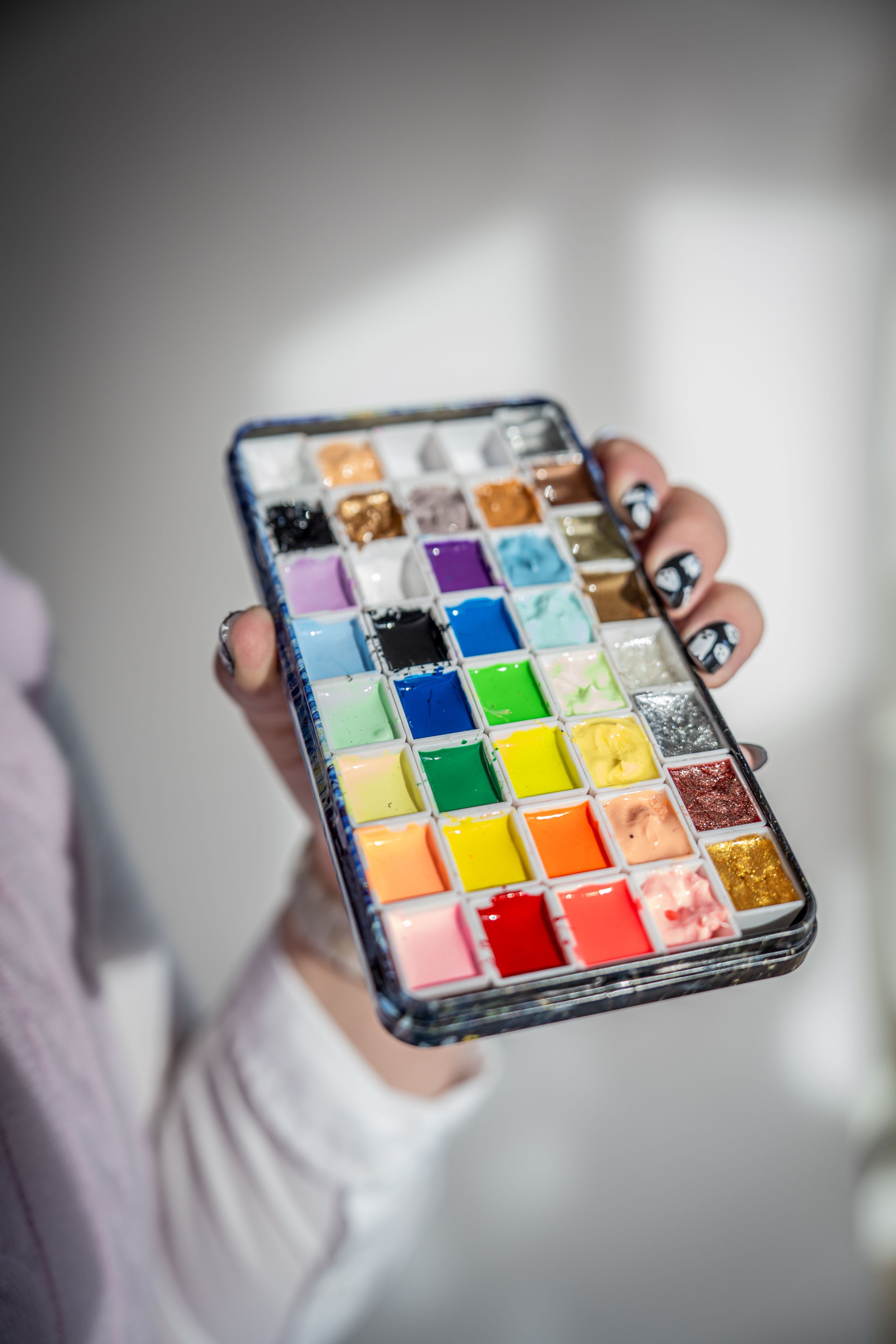 Hand holding a colorful watercolor paint palette with rainbow hues and decorated nails against white background.