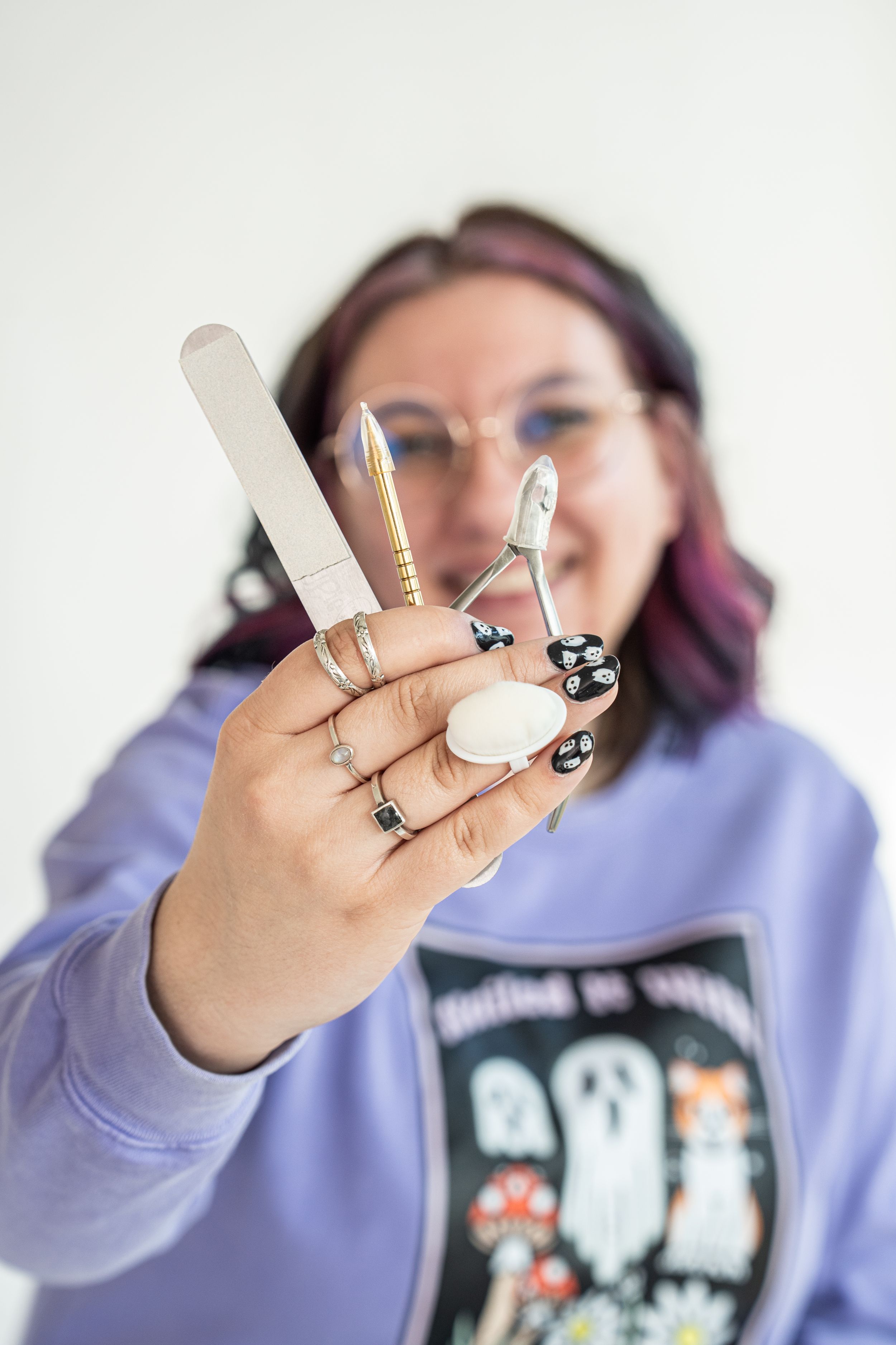 Woman with colorful hair holding nail file and scissors, showcasing black floral nail art, wearing purple hoodie.