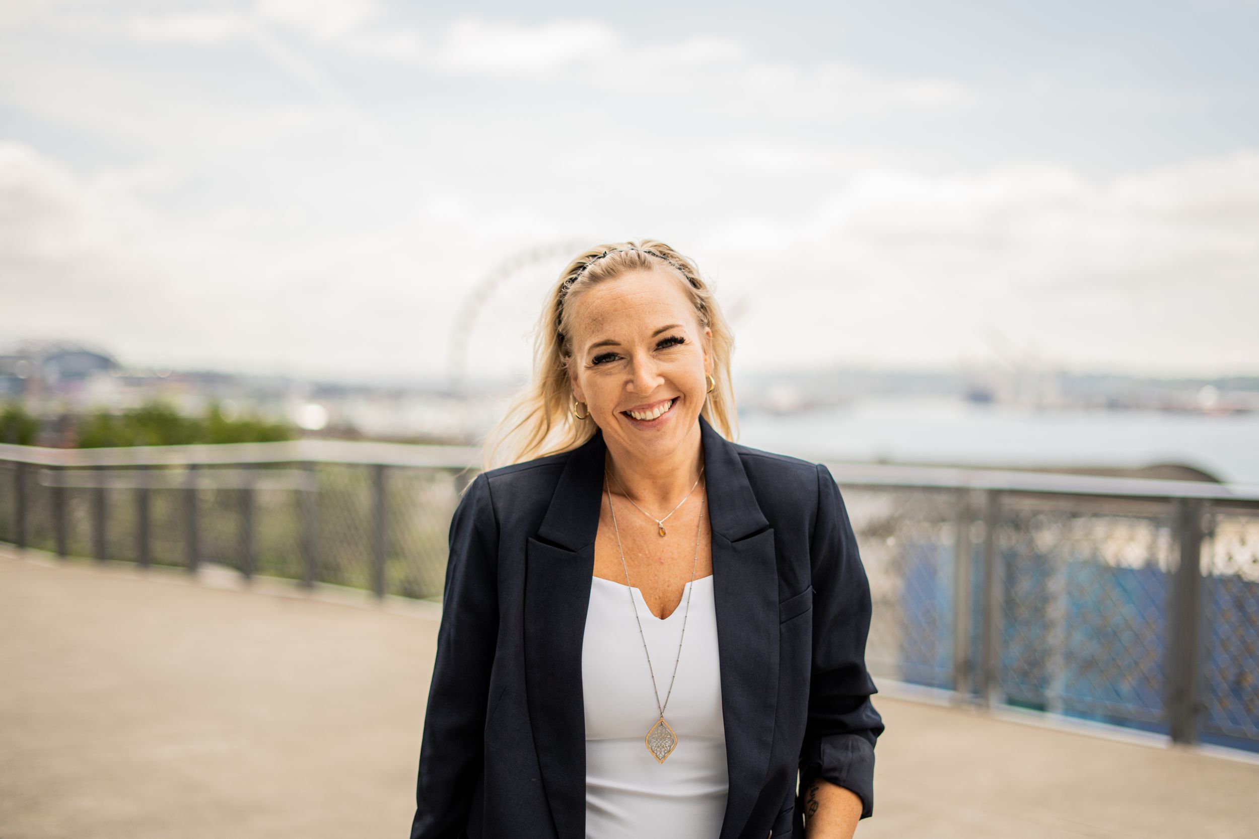 Smiling blonde woman in navy blazer and white top posing outdoors near waterfront with Ferris wheel backdrop.