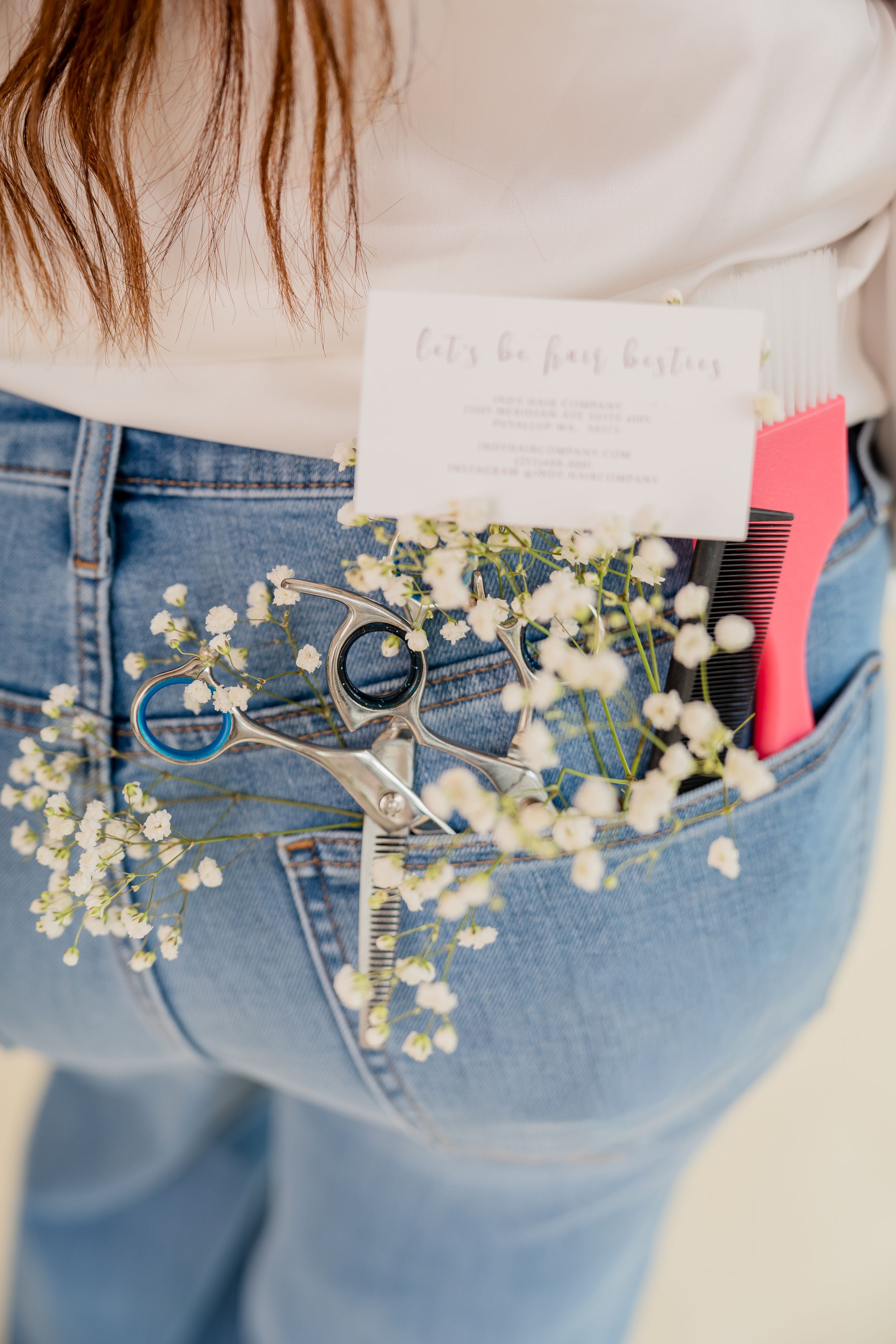 Jeans back pocket holding scissors, comb, baby's breath flowers, and a hair stylist business card.