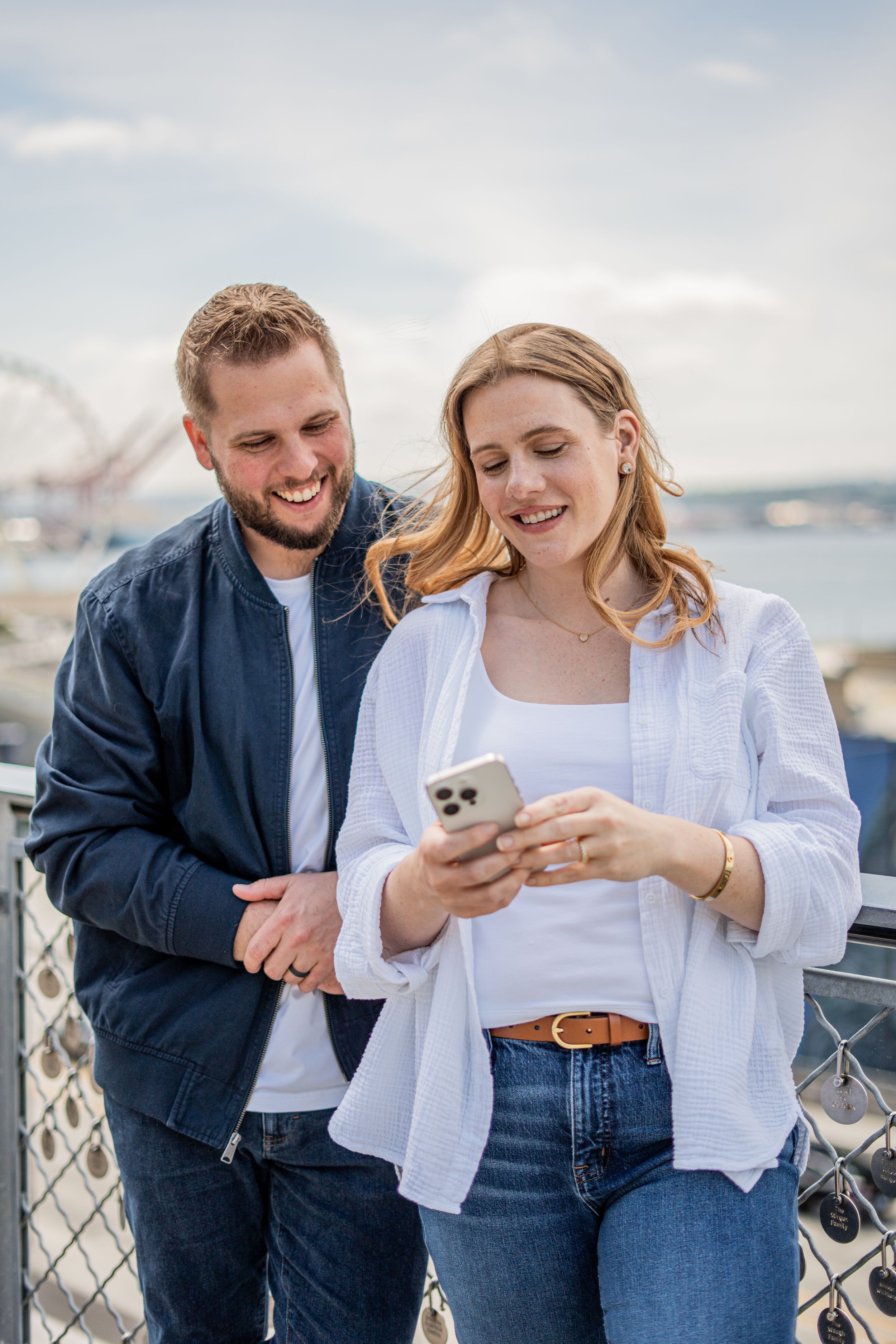 A smiling couple looks at a smartphone together on a waterfront boardwalk with a ferris wheel in the background.