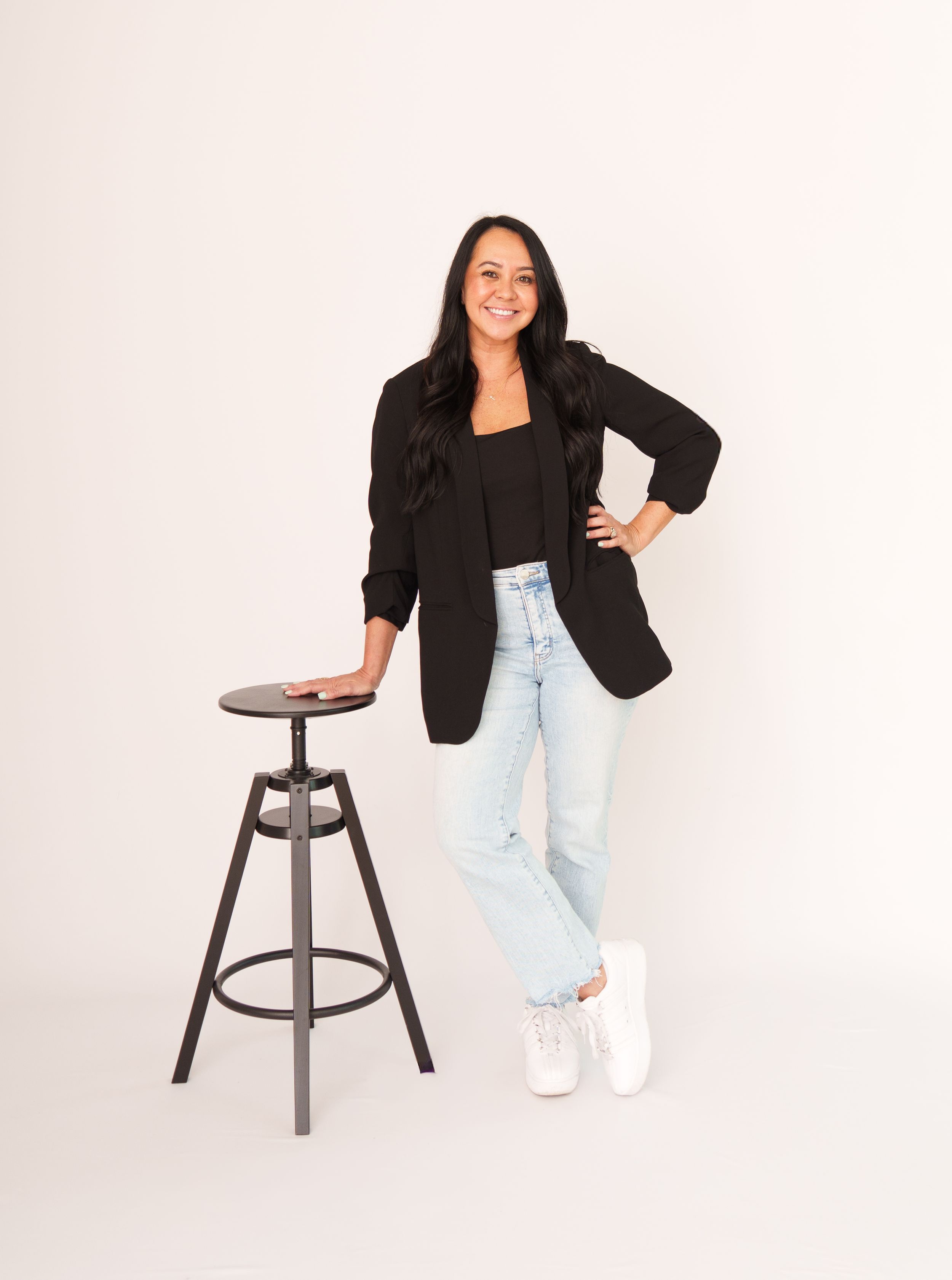 Woman in black blazer and light denim jeans poses casually with industrial stool against white background.