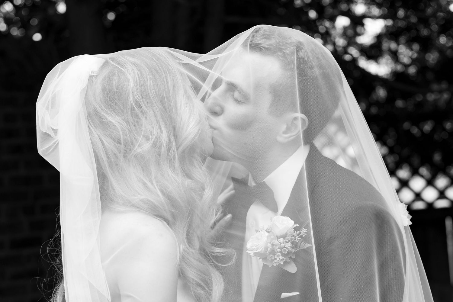 A tender kiss shared between newlyweds under a flowing white veil in an elegant black and white wedding photograph.