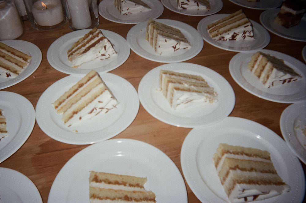 Multiple plates of layered white wedding cake slices arranged on wooden tables. Film Photograph.