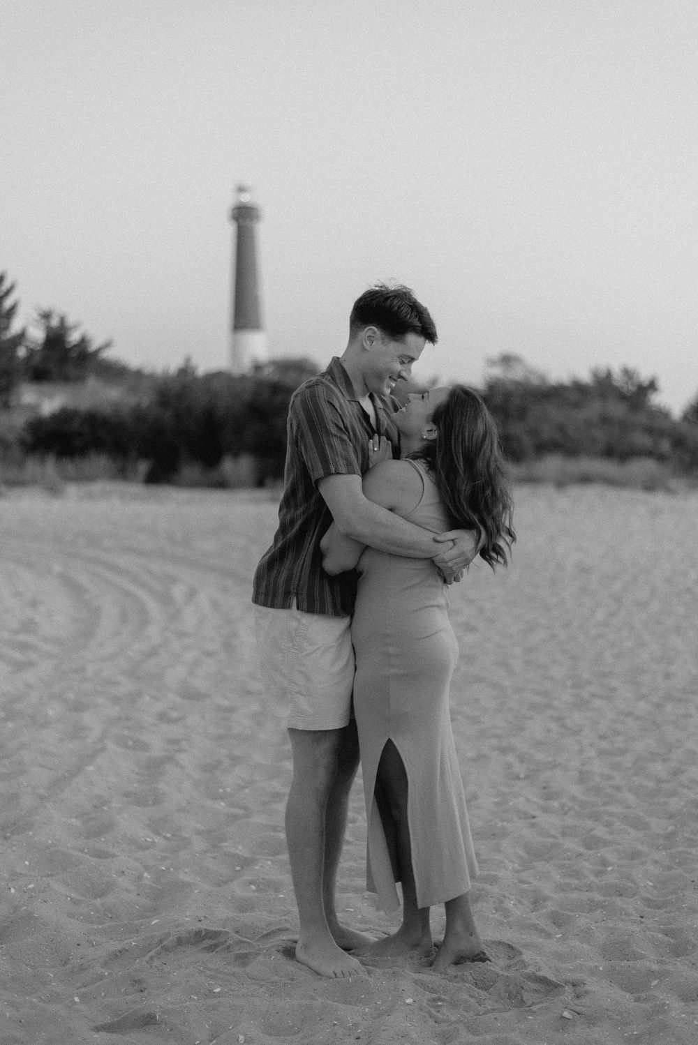 A romantic black and white beach portrait with a lighthouse in the background.