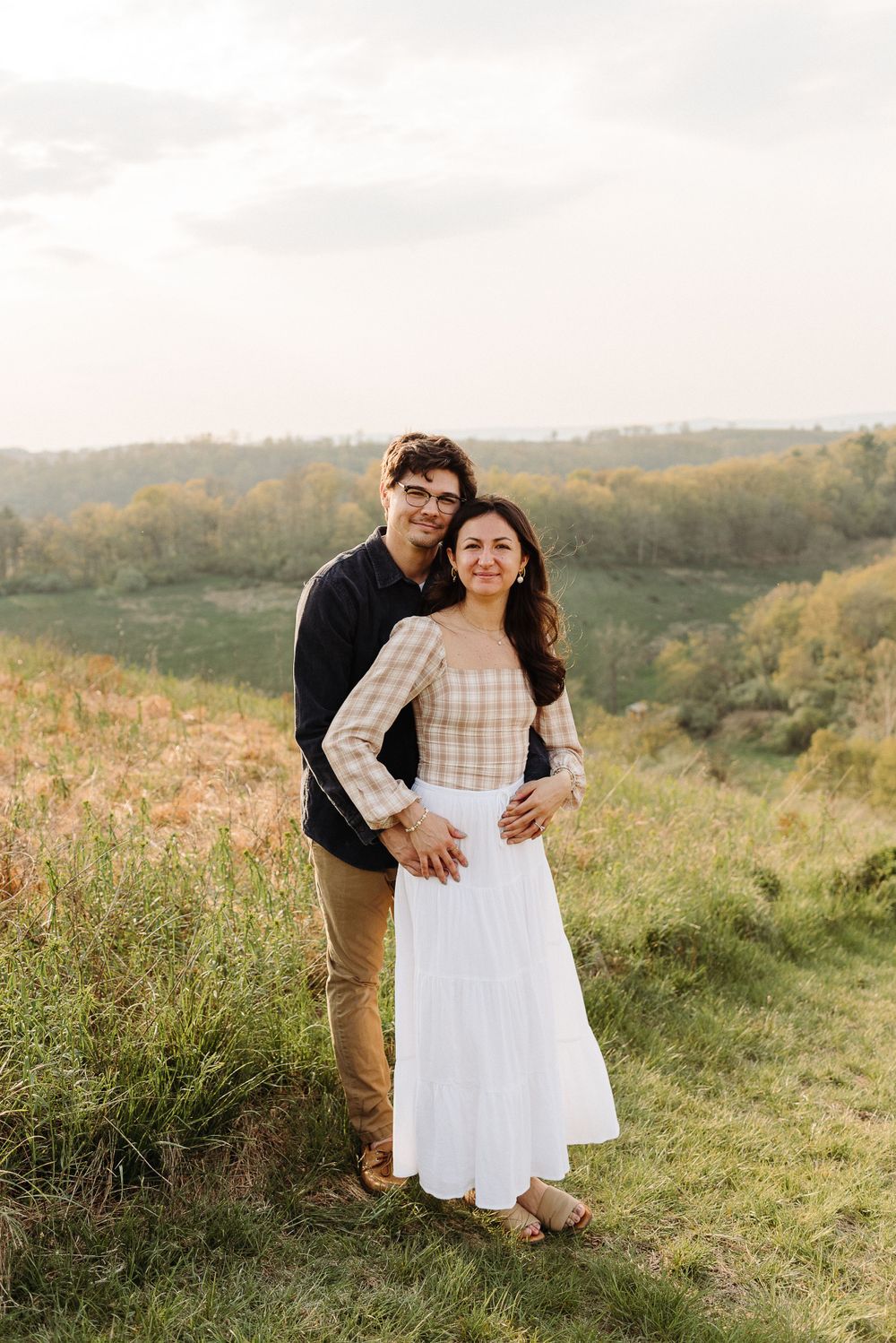 Couple embracing in a sunny meadow with rolling hills in the background.