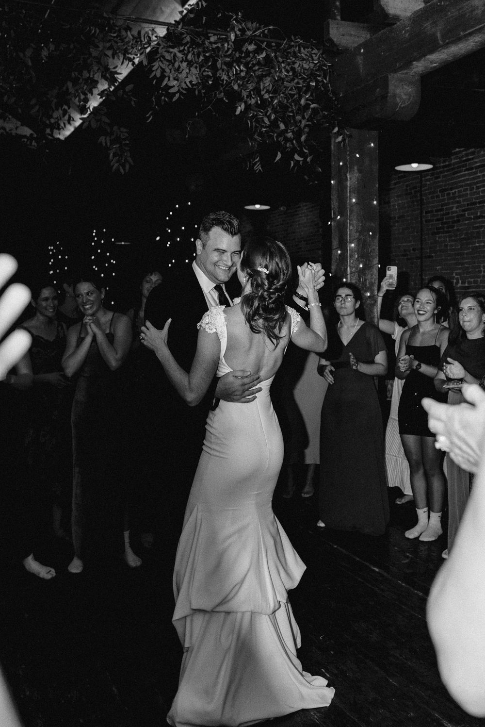 Black and white photo of newlyweds sharing their first dance surrounded by guests.