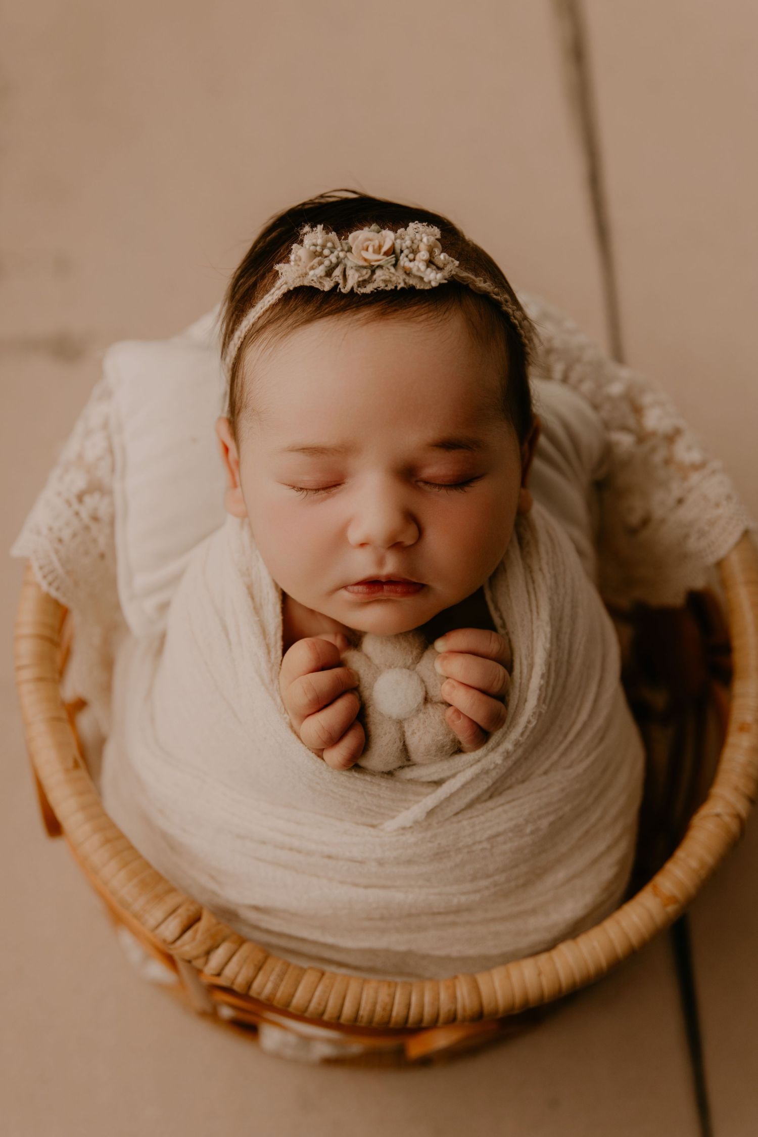 Sleeping newborn baby girl wrapped in white swaddle with floral headband, nestled in a wicker basket.