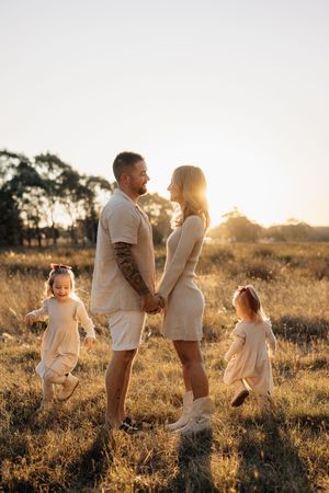 Family in matching white outfits holding hands in golden sunlit field at sunset while children play.