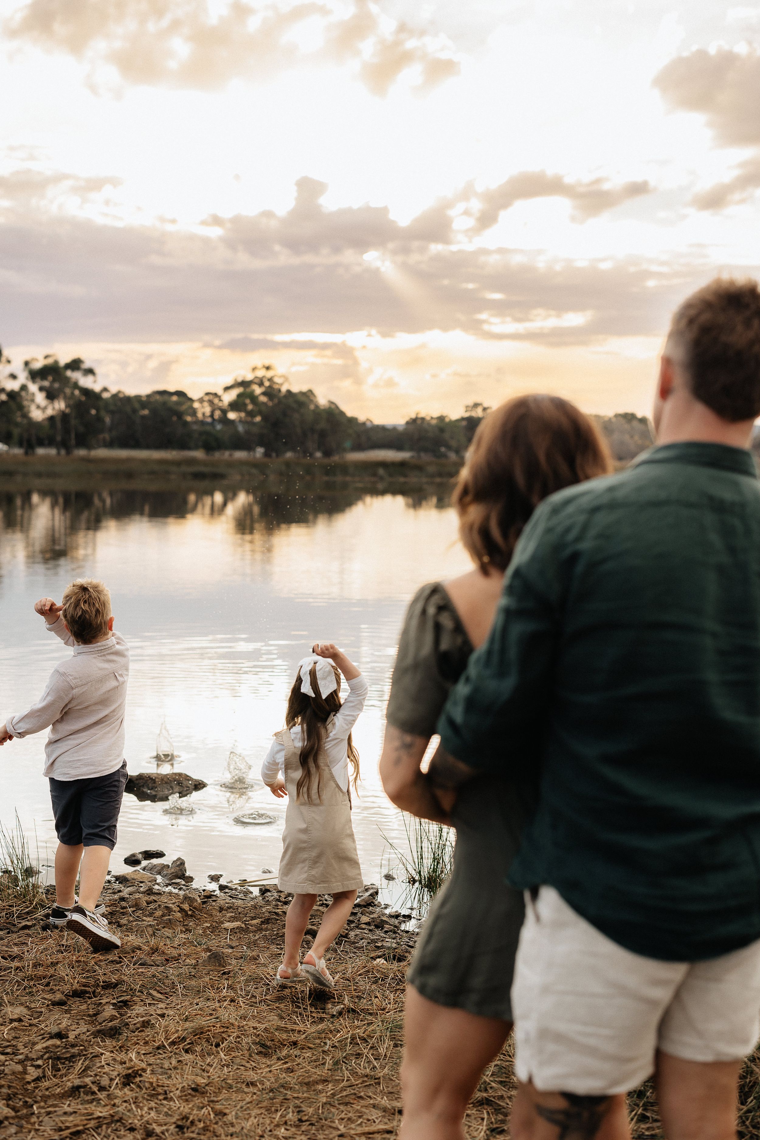 Family enjoying a peaceful sunset by a lake with reflective water and tree-lined shore in golden evening light.