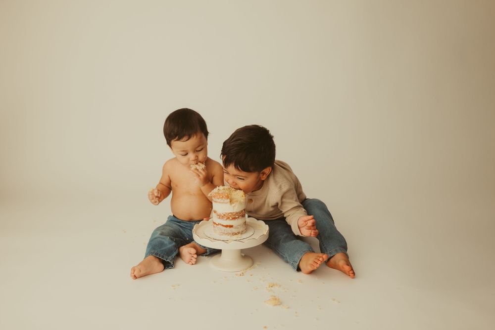 Big brother enjoys baby brother's cake smash cake with him on minimalist backdrop in Clarksville, Tennessee photography studio.