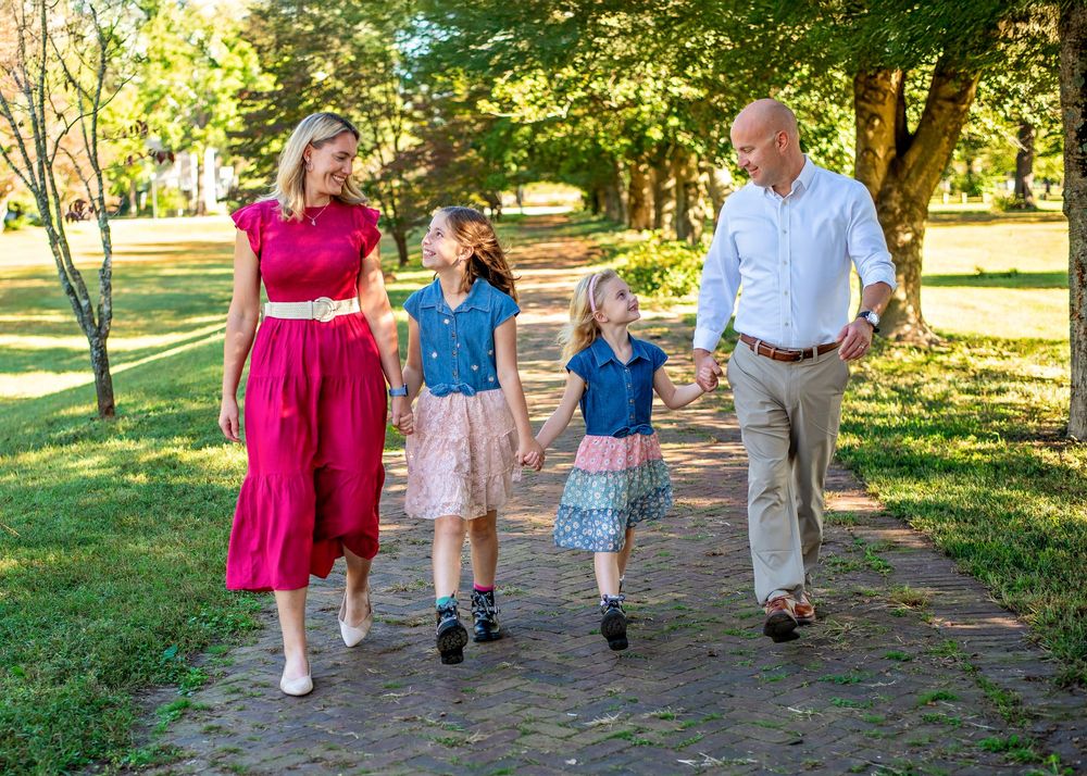 Family members walk together on a tree-lined path in a park during golden hour sunlight.